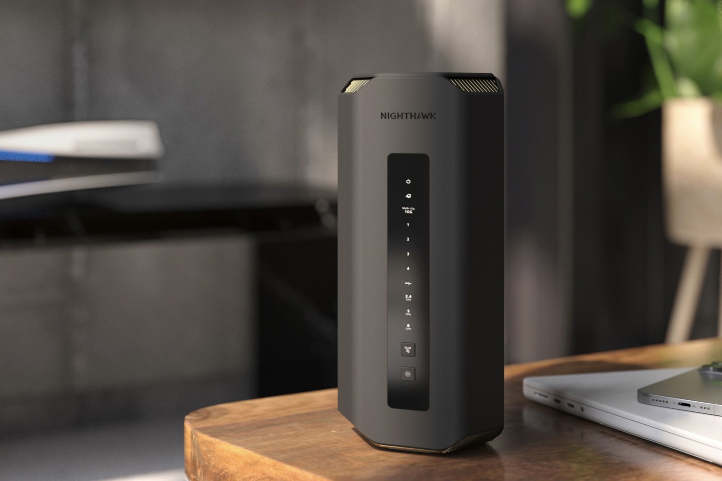 Tower router with a matte black finish on a wooden table, a PS5 is in the distance, and an iPhone and MacBook Pro sits on the table next to the Netgrear nighthawk.